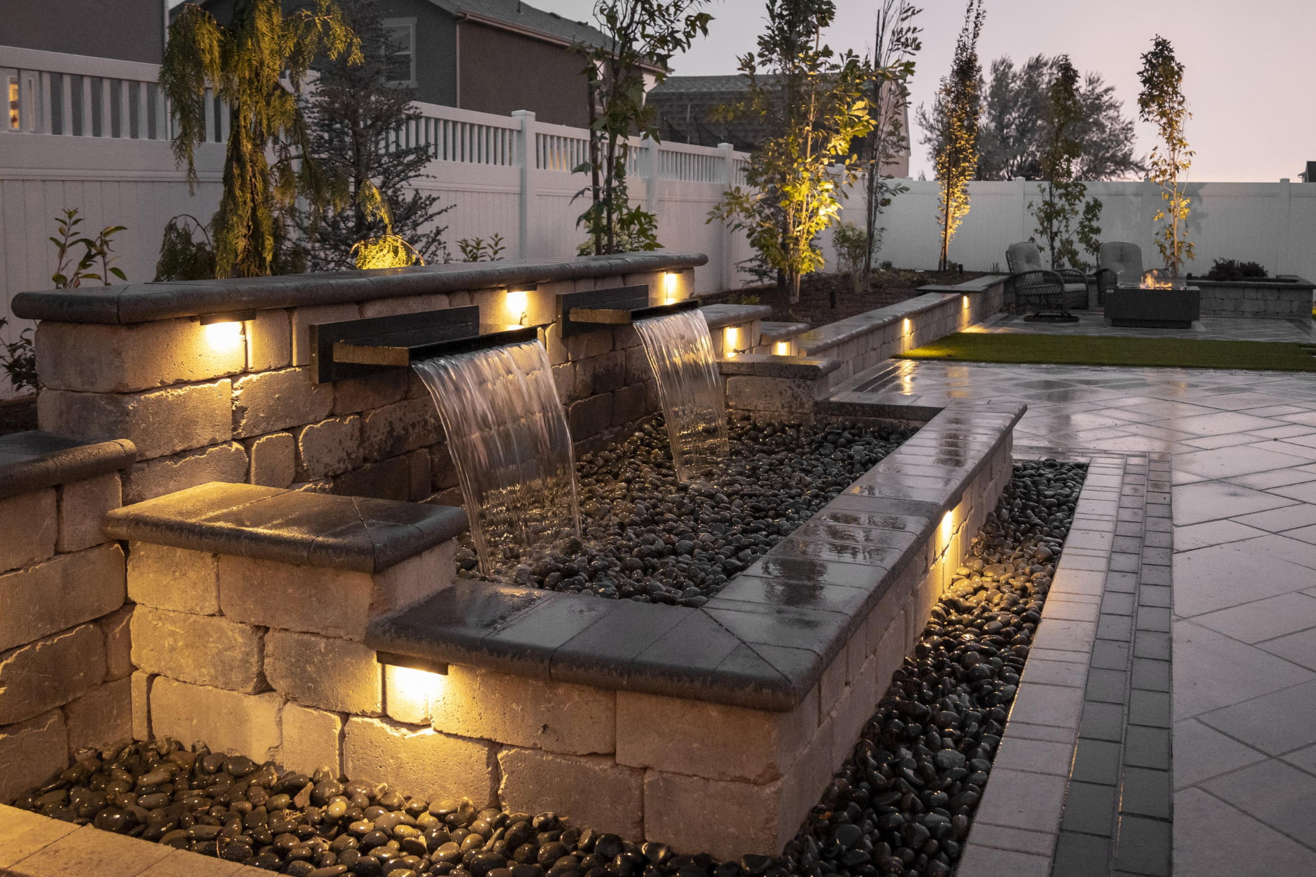 Full water wall run at dusk with wet pavers and a fire table at the far end